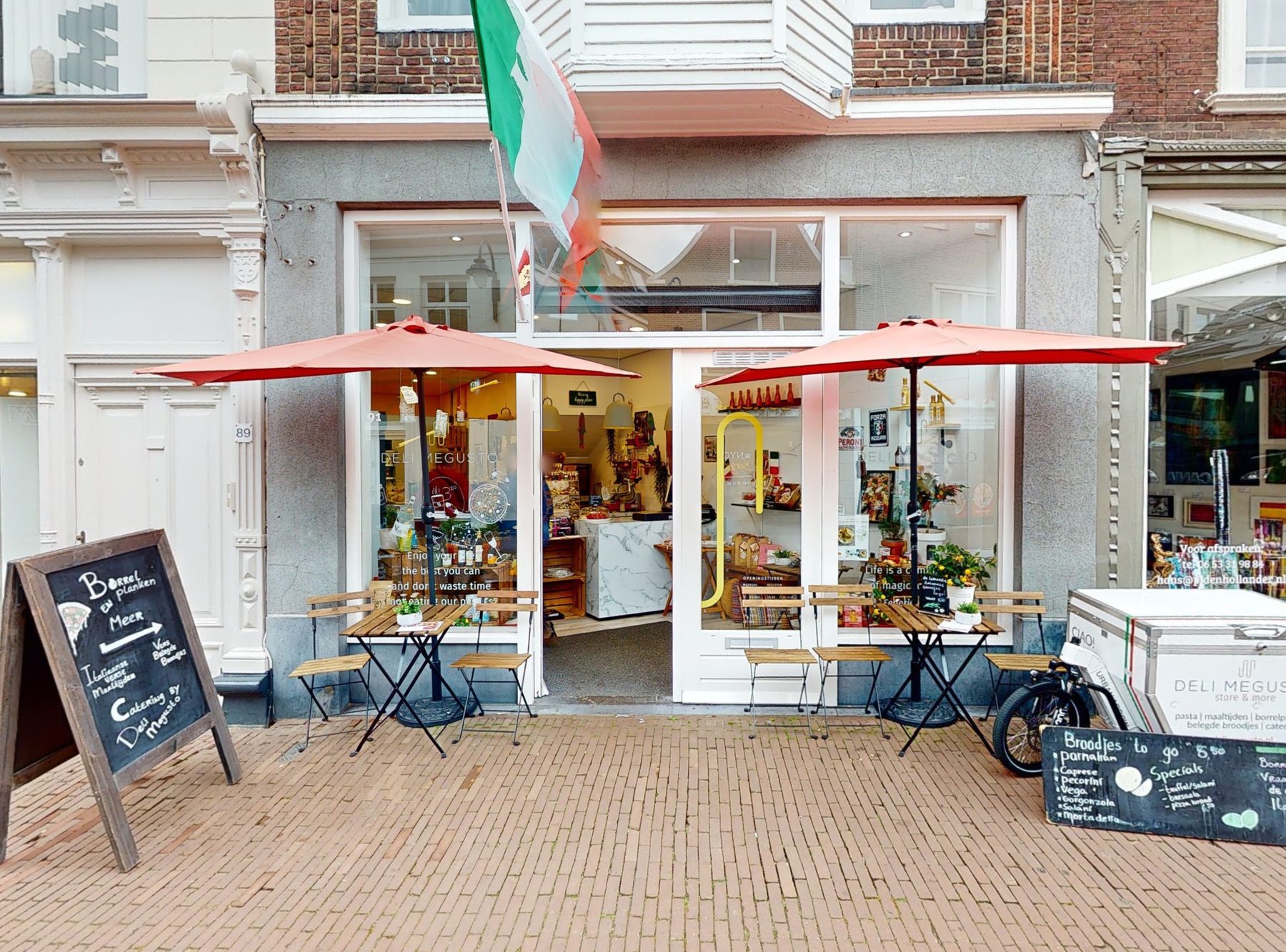 De voorkant van Deli MeGusto aan de Kleiweg 91: Italiaanse vlag, rode parasols, bistrotafeltjes, krijtborden en de MeGusto-bakfiets